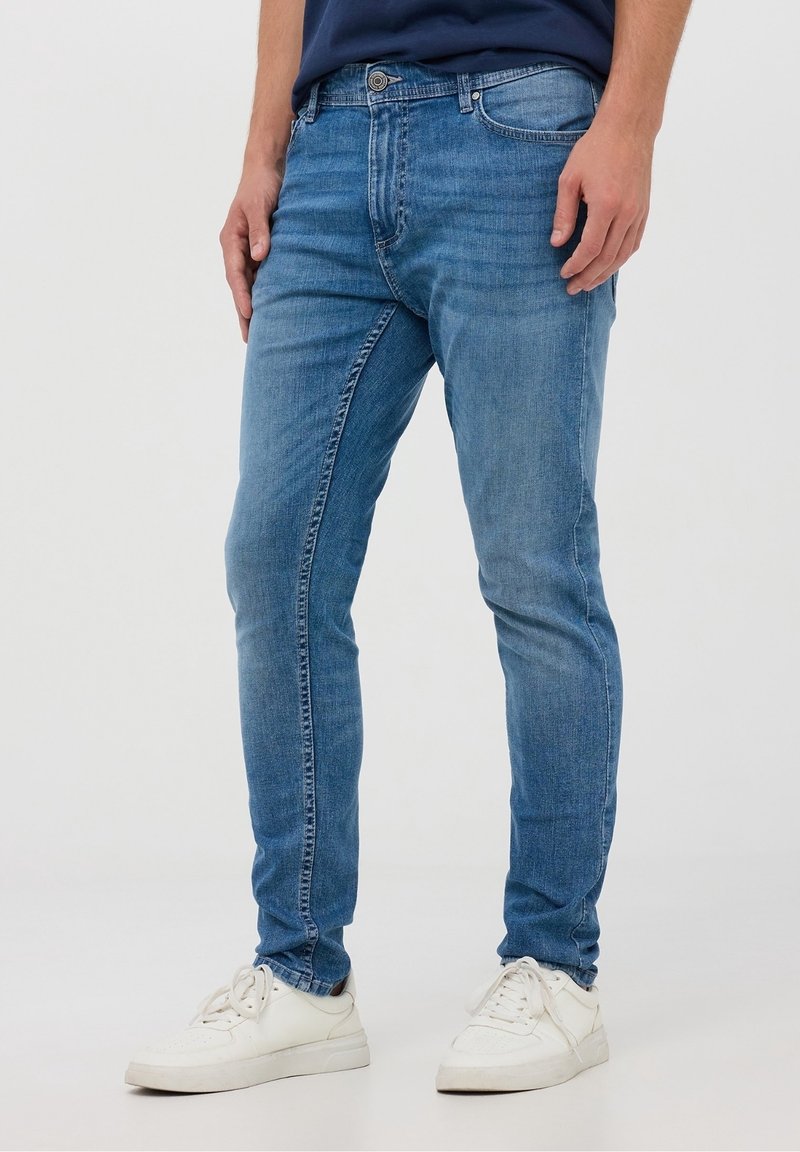 Jeans bleu slim en denim, avec un design classique à cinq poches et un effet légèrement délavé, assortis à des baskets blanches.
