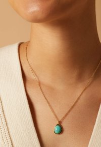 Gouden ketting met een druppelvormige turquoise hanger met een gladde surface en gepolijste afwerking, rustend op een lichte stof.