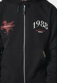 Felpa nera con zip realizzata in misto cotone. Presenta una grafica rossa di un cavallo alato, il testo "1988" e dettagli a motivo check.