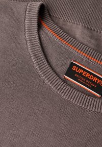 Maglione grigio con collo a girocollo a coste, caratterizzato da una rifinitura arancione a contrasto. Include un'etichetta con il marchio "Superdry" e istruzioni per la cura.