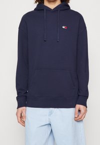 Navy blue pullover hoodie made of soft fabric, featuring a front kangaroo pocket, drawstring hood, and logo patch on the chest.