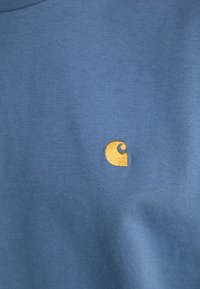 T-shirt in cotone blu con una texture liscia, caratterizzata da un piccolo logo giallo ricamato sul davanti. Design semplice senza ulteriori motivi.