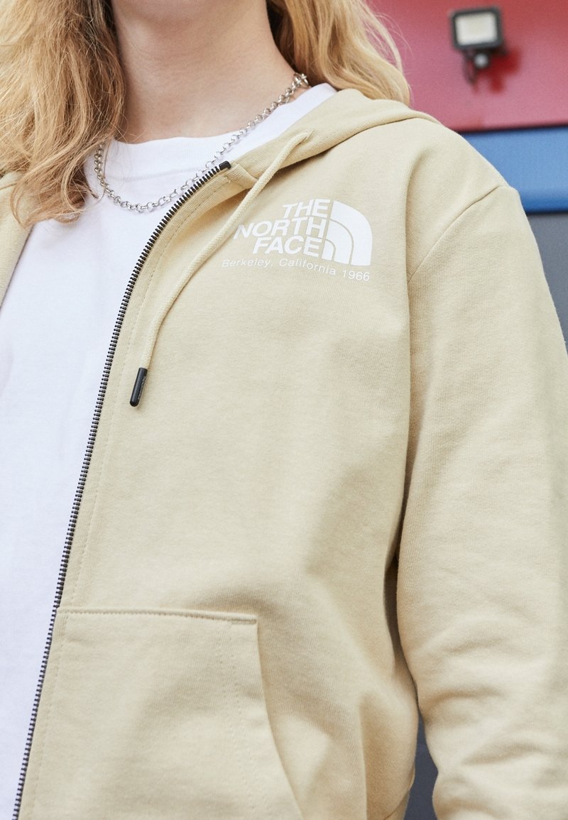 Personne portant un hoodie zippé beige The North Face sur une chemise blanche et un collier chaîne argenté, avec de longs cheveux blonds visibles.