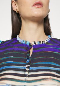 Femme portant un chemisier rayé bleu, violet et beige, avec un col à volants et deux boutons foncés au niveau de l'encolure, et de petites boucles d'oreilles en or.