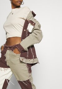 Person wearing a beige cropped top and patchwork, frayed denim jacket and pants in beige and brown tones, with hands in pockets.