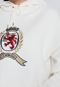 Felpa bianca con cappuccio a cordino, caratterizzata da un significativo emblema di leone rosso circondato da rami d'ulivo e un banner dorato con la scritta "EST. MCMLXXXV."