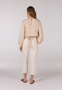 Beige cropped blouse met lange ballonmouwen, strikdetail aan de achterkant en een sleutelgat-halslijn, gecombineerd met witte broek met opgerolde mouwen en zwarte sandalen.