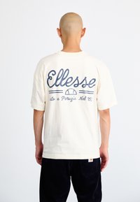 Kreminės spalvos trumpomis rankovėmis marškinėliai su mėlyna "Ellesse" logo ir tekstu nugaroje. Kasdienis dizainas, minkšta medžiaga ir laisvas pasiuvimas.