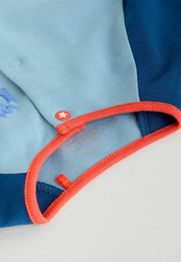 Cotton shirt featuring blue and light blue panels. Orange ribbed collar with a red snap button adorned with a white star. Soft texture.