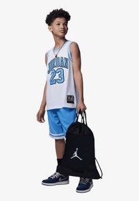 Conjunto de basquetebol Jordan com uma camisola de malha branca com realces azuis, calções azuis a condizer e uma mala de cordão preta com um logótipo.