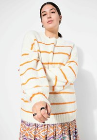 Cremefarbener Pullover mit orangefarbenen horizontalen Streifen, weiten Ärmeln und Strickstruktur. Kombiniert mit einem gemusterten Rock in lebhaften Farben.