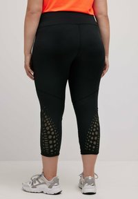 Zwarte capri leggings met een brede tailleband, voorzien van een getextureerd paneel met ronde uitsparingen aan de zijkanten. Achteraan is er gedetailleerde stiksels te zien.
