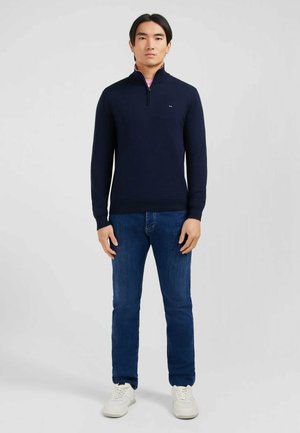 Pull à col zippé marine en matériau doux, avec des poignets et un ourlet côtelés. Associé à un jean bleu et des baskets blanches. Design simple.