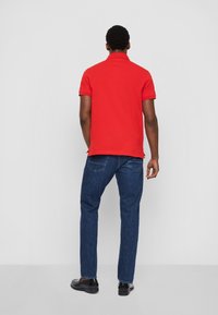 T-shirt polo rouge avec col, manches courtes et fentes sur les côtés, associé à un jean en denim bleu foncé et des chaussures noires, vu de dos.