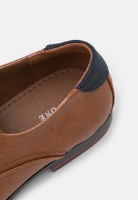 Brązowe skórzane buty z akcentem z ciemnoniebieskiego zamszu na pięcie oraz widocznym szwem. Gładka tekstura, zaokrąglony kształt i solidna podeszwa.
