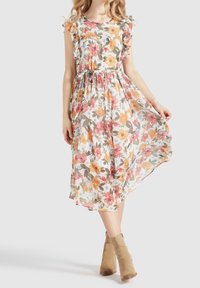 Frau, die ein florales Midikleid mit Rüschenärmeln und einem Bindeband in der Taille trägt, kombiniert mit beigen Wildleder-Stiefeletten, stehend vor einem einfarbigen Hintergrund.