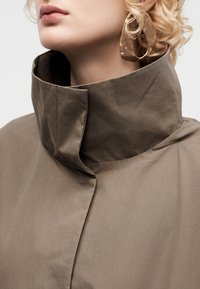 Κοντινό πλάνο ενός ατόμου που φοράει παλτό ταμπά με ψηλό γιακά και μεγάλα, υφασμένα χρυσά σκουλαρίκια κρίκους, με εμφανή τα χείλη και μέρος των σγουρών ξανθών μαλλιών.