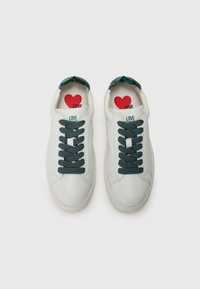 Baltas ādas sporta apavi ar tumši zaļām šnūrēm un strukturētu zaļu iekšējo oderi. Iekšpusē ir sarkans sirds grafikas attēls.