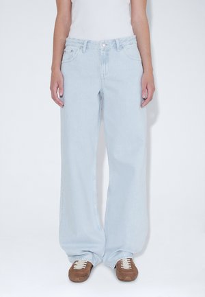 Personne portant un jean large bleu clair taille haute et des baskets marron, debout sur un fond blanc uni.