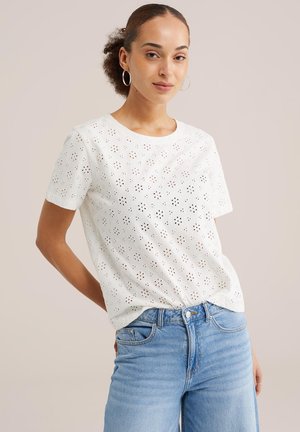 Fiatal nő göndör hajjal, ezüst karika fülbevalókat visel, fehér hímzett rövid ujjú felsőt és világoskék, magas derekú farmert, hátratett kézzel áll.