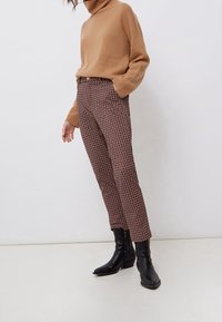 Femme portant un pull à col roulé camel, un pantalon marron à motifs et des bottines noires, debout devant un fond blanc uni.