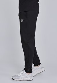 Crne trenirke slim fit kroja, s malim bijelim logom. Tkanina djeluje glatko i rastezljivo, s patentnim zatvaračima na gležnjevima.
