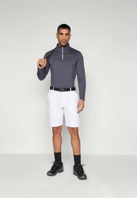 Férfi áll, vállán tartva a golfütőt, szürke hosszú ujjú pólót, fehér sortot, fekete övet, fekete zoknit és fekete golfcipőt viselve.