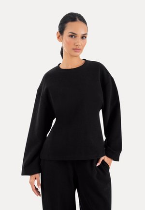 Kobieta o ciemnych włosach związanych wstecz, ubrana w luźną czarną bluzkę z długim rękawem i pasujące czarne spodnie, stoi na tle jednolitego tła.