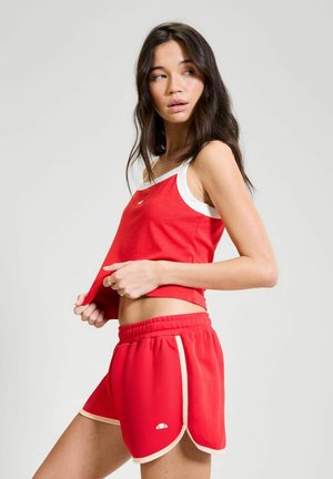 Vrouw in rood sportief tanktop en bijpassende korte broek, kijkt naar links terwijl ze haar shirt iets omhoog tilt, tegen een effen lichte achtergrond.