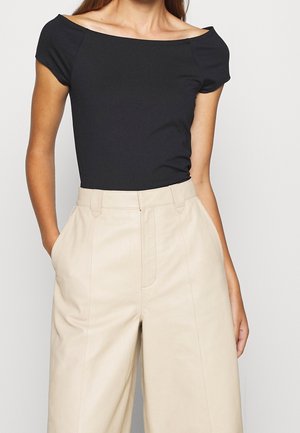 Woman wearing a fitted black off-shoulder top and high-waisted wide-leg beige pants with hands in pockets against a neutral background.