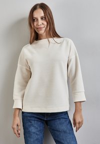 Světlý béžový top s texturovaným žebrovaným designem, širokým lodním výstřihem a tříčtvrtečními rukávy, doplněný modrými džíny.