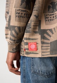 Beige sweatshirt featuring black abstract patterns, ribbed cuffs, and a red logo tag near the hem. Pair with blue denim jeans.