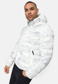 Doudoune blanche avec un motif gris marbré, dotée d'une fermeture éclair, de poches latérales et d'une capuche. Fabriquée en matériau texturé.