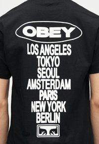 Μαύρο T-shirt με το κείμενο «OBEY» μέσα σε οβάλ, ακολουθούμενο από ονόματα πόλεων: Λος Άντζελες, Τόκιο, Σεούλ, Άμστερνταμ, Παρίσι, Νέα Υόρκη, Βερολίνο, και ένα λογότυπο με στυλιζαρισμένο πρόσωπο.