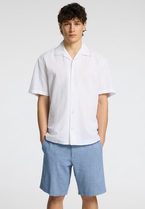 Camicetta bianca in lino a maniche corte con colletto, chiusura frontale a bottoni e pantaloni corti azzurri con un motivo testurizzato. Abbigliamento estivo informale.
