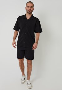 Schwarzes kurzärmeliges Hemd mit Knöpfen, kombiniert mit schwarzen Shorts. Das Material scheint strukturiert zu sein. Zubehör sind weiße Sneaker mit schwarzen Akzenten.