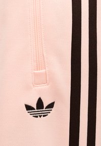 Κοντινό πλάνο ροζ υφάσματος με μαύρο λογότυπο Adidas trefoil και δύο κάθετες μαύρες ρίγες δίπλα σε τσέπη με φερμουάρ.