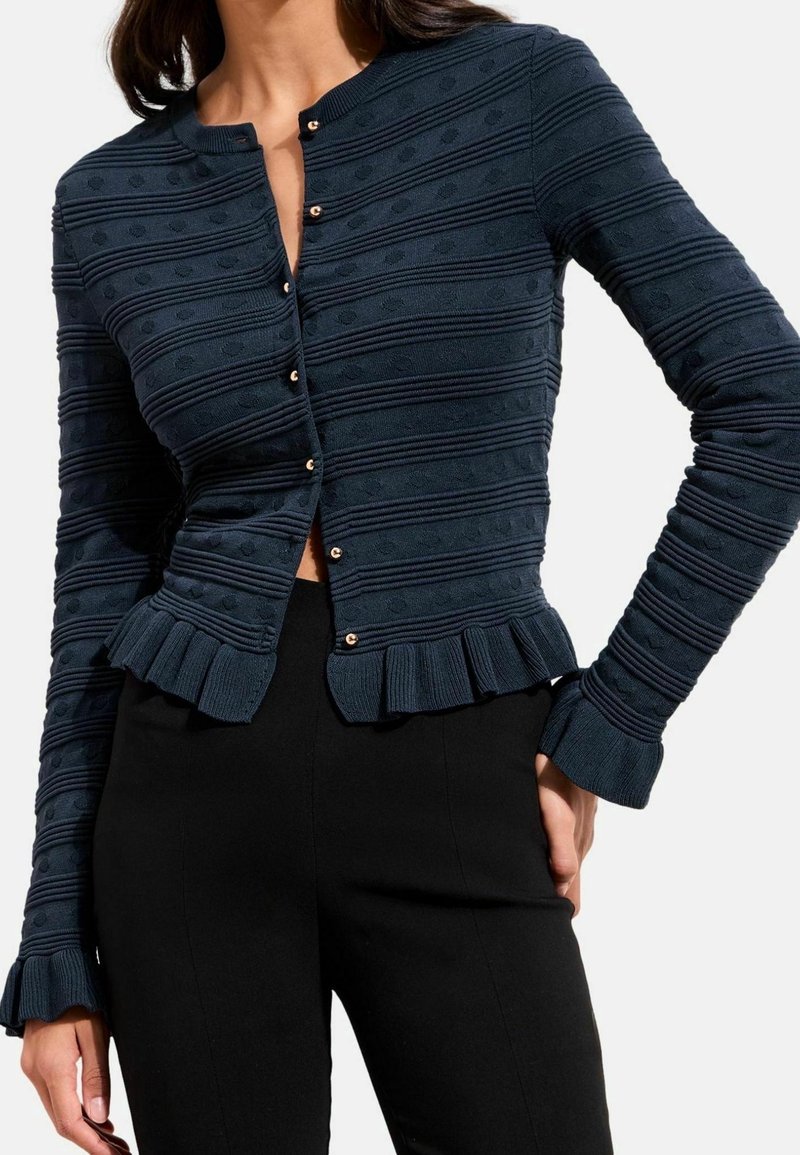 Femme portant un cardigan en maille texturée bleu marine avec de petits boutons, des rayures horizontales et un ourlet à volants, associé à un pantalon noir.