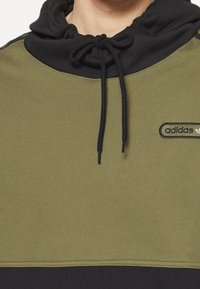 Olivgrön sweatshirt med svart huva och dragsko, med en broderad adidas-logotyp på bröstet. Mjukt material och avslappnad passform.