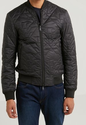 Hombre con chaqueta bomber acolchada negra con cremallera frontal sobre vaqueros azul oscuro, de pie frente a un fondo claro y liso.