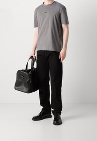 Man med grå BOSS T-shirt, svarta jeans, polerade svarta skor, som håller en stor svart läderväska mot en helt vit bakgrund.