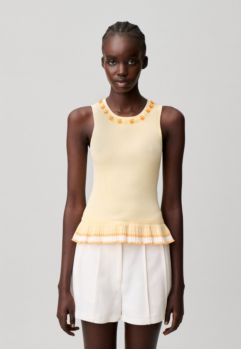 Mujer con una camiseta sin mangas beige con detalles de flores naranjas en el escote y dobladillo fruncido, combinada con pantalones cortos blancos de talle alto.