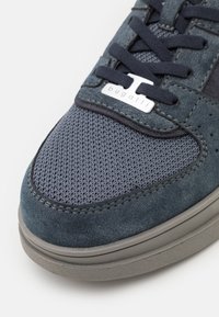 Gros plan sur une basket bleu foncé en suède et mesh, lacets bleu marine, semelle grise, et plaque argentée "bugatti" sur la languette.