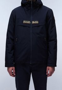 Granatowa wodoodporna kurtka z zamkiem błyskawicznym, regulowanym kapturem i naszywką z logo na klatce piersiowej. Posiada dwie boczne kieszenie na zamek.