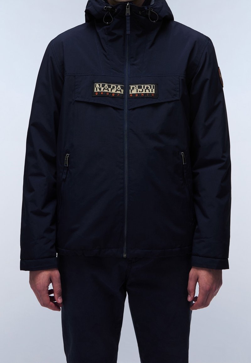 Navyblå vandtæt jakke med lynlåslukning, justerbar hætte og logo-patch på brystet. Har to sidelommer med lynlås.