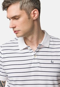 Biała koszulka polo w granatowe poziome paski, z dwoma guzikami i małym logo na piersi. Teksturowana tkanina z mieszanki bawełny.
