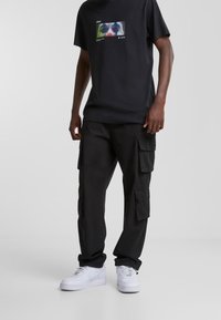Czarny bawełniany t-shirt z grafiką na przodzie, zestawiony z czarnymi spodniami typu cargo z wieloma kieszeniami oraz białymi trampkami. Prosty krój i design.