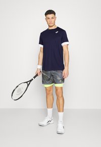 Camiseta deportiva navy con puños blancos, pantalones cortos grises de patrón con ribete verde neón, sosteniendo una raqueta de tenis, usando zapatos y calcetines blancos.