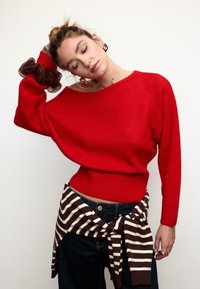 Roter Strickpullover mit geripptem Saum und schulterfreiem Design, kombiniert mit dunkler Jeans und einem braun-weiß gestreiften Strickstoff, der in der Taille gebunden ist.