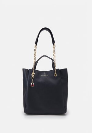 Sac fourre-tout en cuir noir avec des anses en chaîne dorée, de petites poignées, et un logo Tommy Hilfiger avec un charm sur le devant.